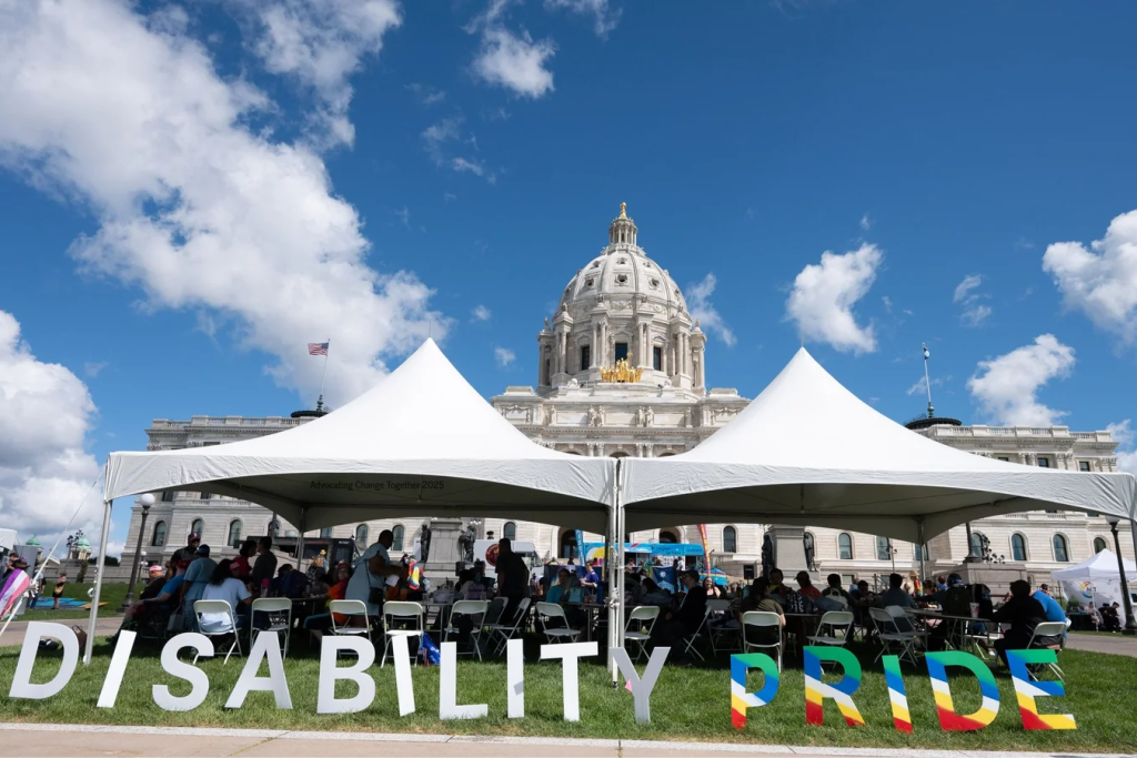 The image depicts a large white tent set up on a lawn in front of the domed state capitol building. Large letters in the foreground spell "DISABILITY PRIDE" in white and rainbow colors.