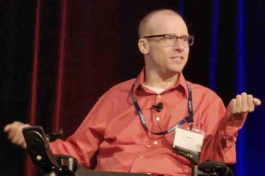 A man in a red shirt speaks from a wheelchair at a conference, sharing insights on disability and advocacy.