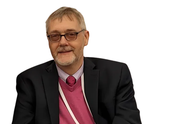 A smiling man in a suit with a pink sweater vest, glasses, and a lanyard, seated against a plain background.