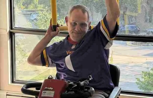 A man in a Vikings jersey exercises using a mobility device, smiling while lifting handles near a sunny window.