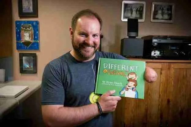 Author Ryan Haack smiles while holding his children's book "Different is Awesome!" in a cozy, art-filled room.