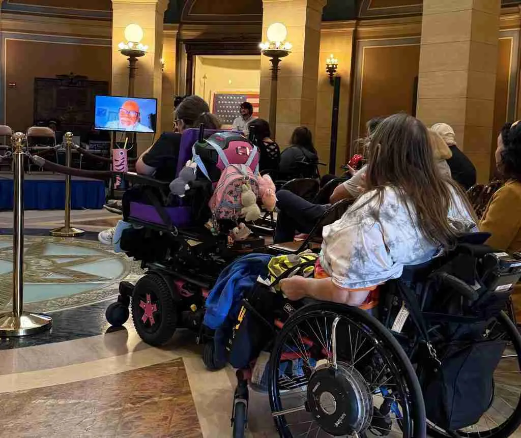 Attendees in wheelchairs participate in a meeting, with a speaker visible on a screen in a formal setting.